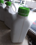 trzy pięciolitrowe plastikowe białe pojemniki z zielonymi nakrętkami stoją na parapecie okna; przez okno widać ciężarówki stojące na przejściu granicznym