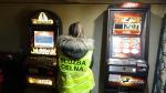 Funkcjonariusza celno - skarbowa stojąca tyłem pomiędzy dwoma automatami do gier hazardowych. Automaty włączone, na wyświetlaczach kolorowe znaki.