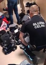 Funkcjonariusz Służby Celno-Skarbowej kucający przy zabezpieczonej odzieży. Na podłodze porozkładane bluzy, czapki itp.