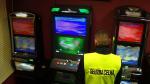Funkcjonariusz Służby Celno-Skarbowej stoi przed trzema automatami do gier hazardowych. Automaty są włączone, działają.
