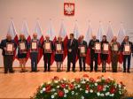 Wspólne zdjęcie wszystkich nagrodzonych dyplomami uznania z medalem wręczonych przez Wojewodę Lubelskiego. Uczestnicy stoją na tle flag Polski. Na pierwszym planie widoczna kompozycja kwiatowa.