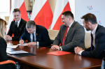 Uczestnicy spotkania siedzą przy stole konferencyjnym. Starosta Powiatu Radzyńskiego podpisuje dokumenty. W tle widoczne flagi państwowe i ścianka z napisami KAS.